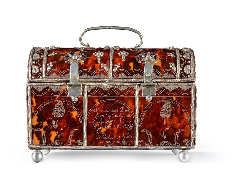 Back of casket made of tortoiseshell with a silver lock and handle, silver decoration, and silver ball feet. Silver decoration takes the form of seashells and abstract geometric patterns. Encised message on central panel in Spanish detailing the maker and date. Photographed on a white background.