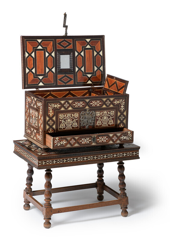 Ornate, rectangular chest in wood on a matching four-legged stand with tortoiseshell, mother-of-pearl, and ivory inlay in floral and geometric pattersn and iron fittings. The lid of the chest is open, revealing a geometric inlay interior, a mirror on the top of the lid, and an open interior flap. A drawer on the bottom of the chest has been pulled out a few inches. The objects are photographed on a white background.