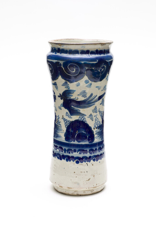 Tin-glazed ceramic jar that flares at the top and the bottom. Decorated in blue-on-white glaze with a bird flying over a landscape with clouds in the sky. Photographed on a white background.