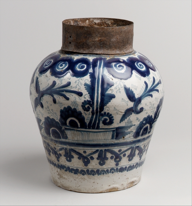 Tin-glazed ceramic jar that flares at the top and the bottom. Decorated in blue-on-white glaze with a bird flying over a landscape with clouds in the sky and a pattern of stylized Arabic letters. Photographed on a white background.