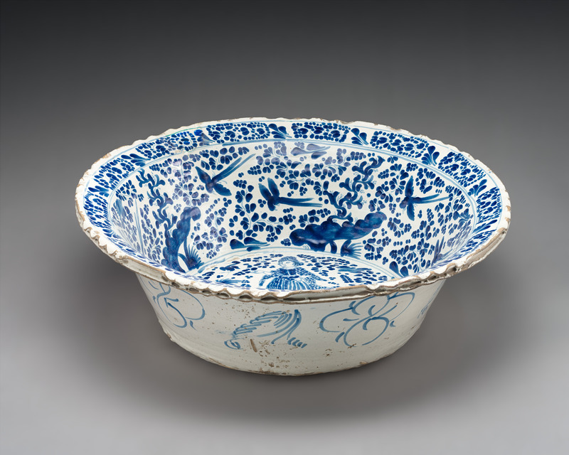 Tin-glazed ceramic basin with an extended lip seen slightly from above and photographed on a neutral gray background. Decorated with blue-on-white glaze with birds and flowers on the sides of the interior, a woman on the bottom of the base, and minimal floral decoration on the exterior sides.