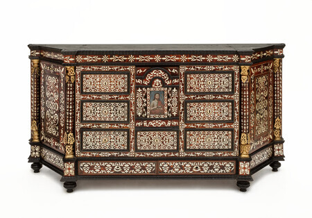 Low, rhombus shaped wooden cabinet with tortoiseshell and mother-of-pearl inlay in abstract floral pattern. In center of the front panel is a painting of a bearded man with long brown hair wearing a gray hair shirt and a red tunic. The man has a halo, carries a crucifix, a bible, and a sheep figurine. The object is photographed on a white background. 