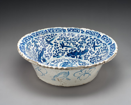 Tin-glazed ceramic basin with an extended lip seen slightly from above and photographed on a neutral gray background. Decorated with blue-on-white glaze with birds and flowers on the sides of the interior, a woman on the bottom of the base, and minimal floral decoration on the exterior sides.